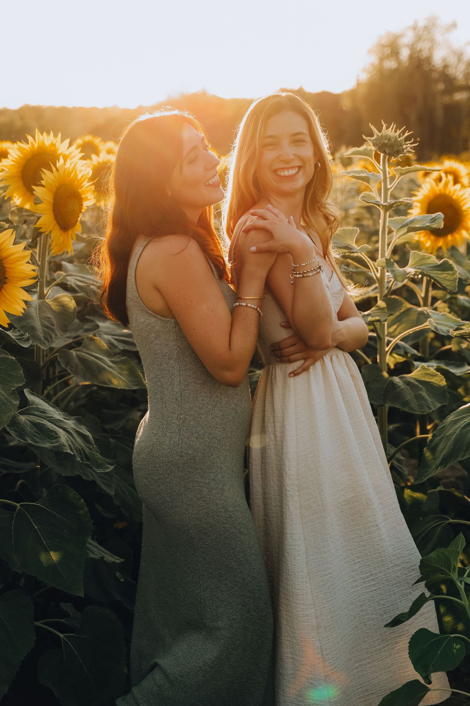 Two smiling women in summer dresses posing together in sunflower field at sunset.