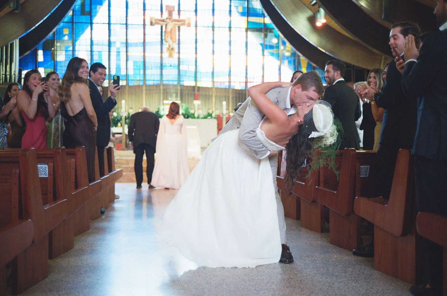 Couple shares a romantic dip kiss in a church aisle with stained glass windows and wooden pews in the background.