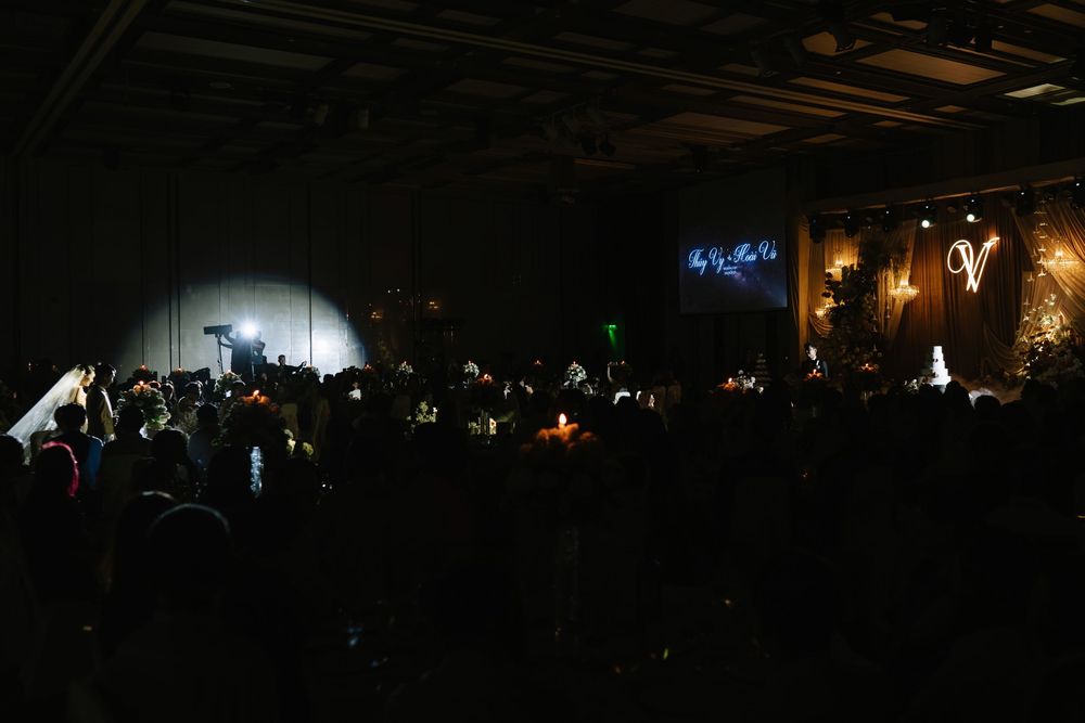 Dark wedding reception venue with dramatic spotlight illuminating a crowded elegant ballroom with decorative monogram display.