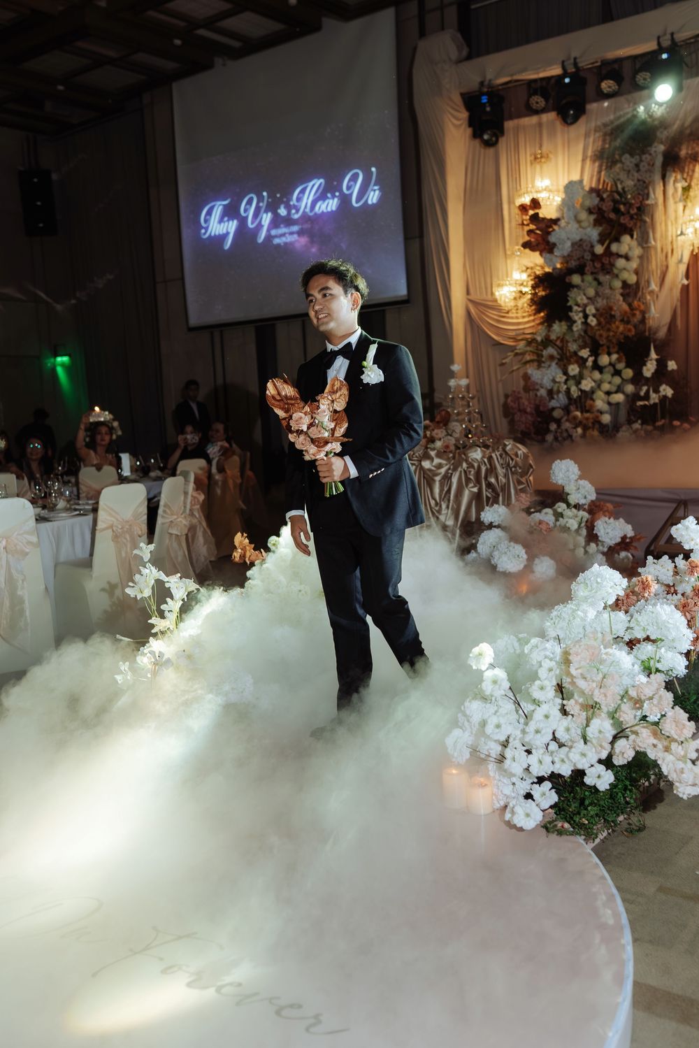 Groom in black tuxedo walks through dry ice fog carrying bouquet at romantic wedding reception entrance.