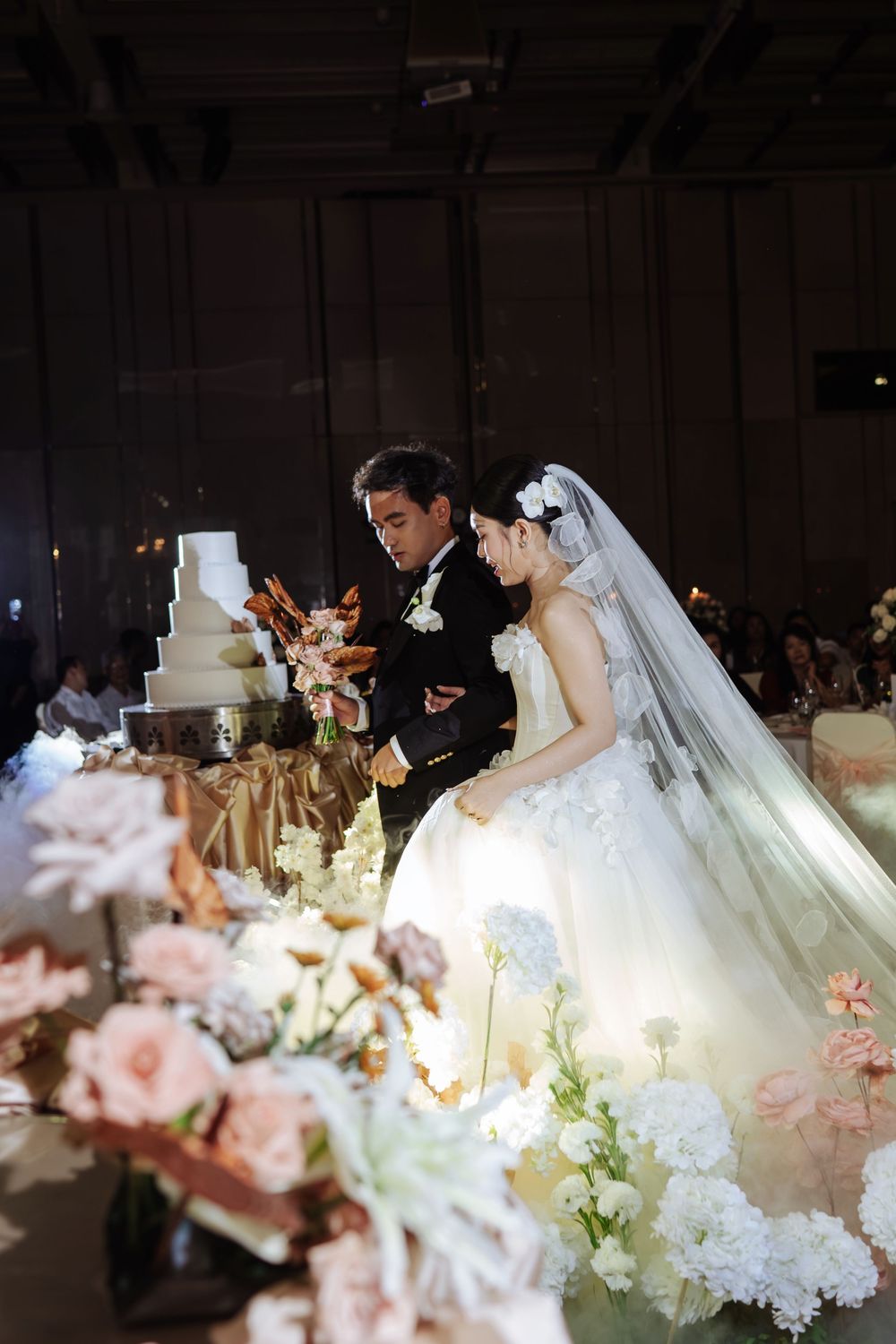 Bride in white gown and groom in tuxedo stand together beside elegant multi-tier wedding cake.