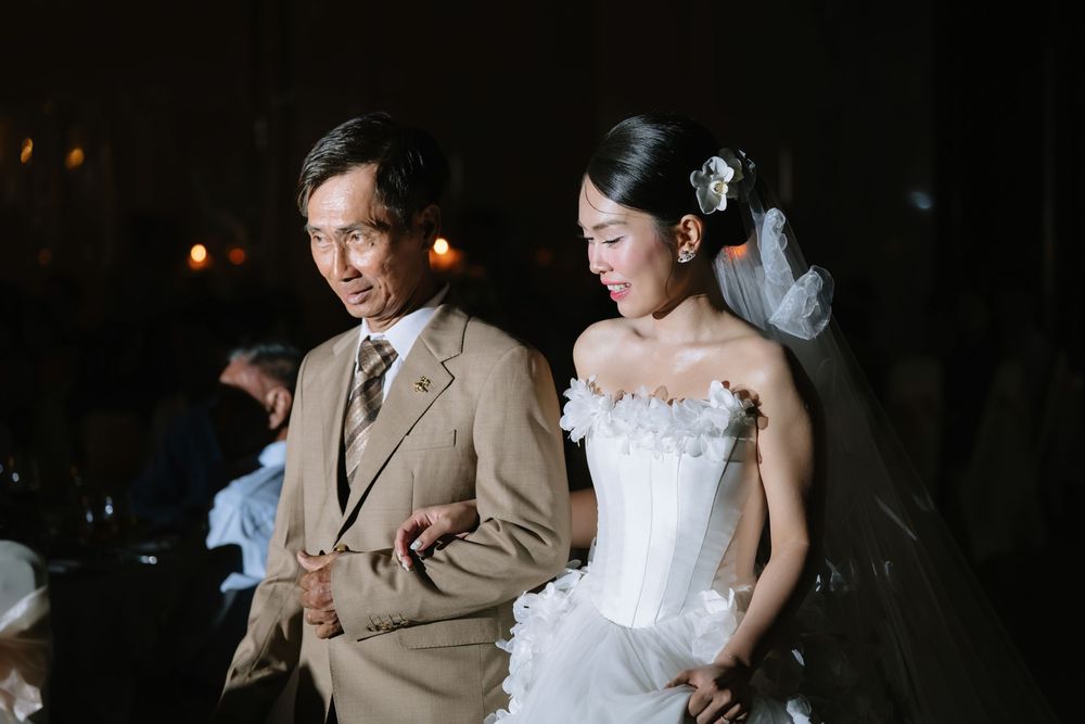 Bride in strapless white wedding gown walks with escort through dimly lit romantic wedding reception.