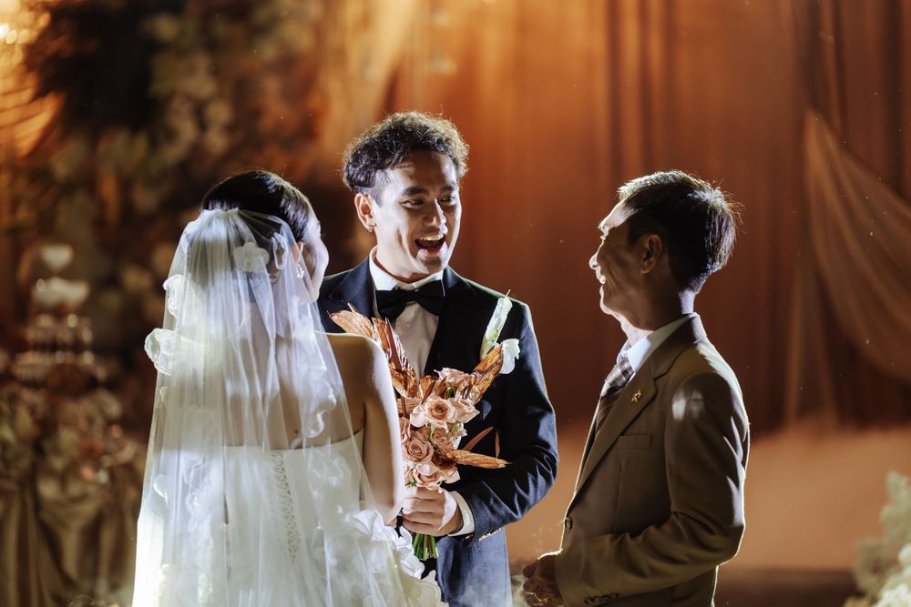 Groom in black tuxedo smiles during wedding ring exchange ceremony with officiant and veiled bride.