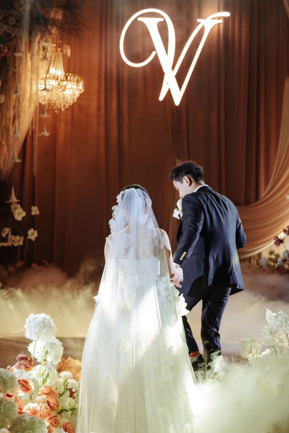 Bride and groom walk hand in hand toward glowing monogram V backdrop surrounded by white florals.
