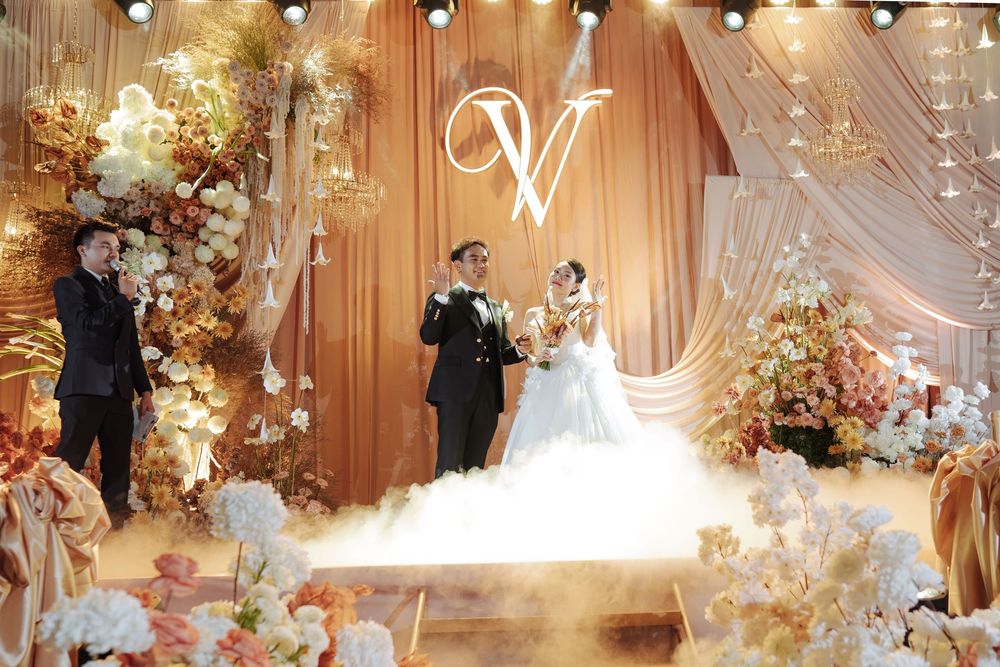 Bride and groom pose before golden draped backdrop with floral arrangements and glowing V monogram.