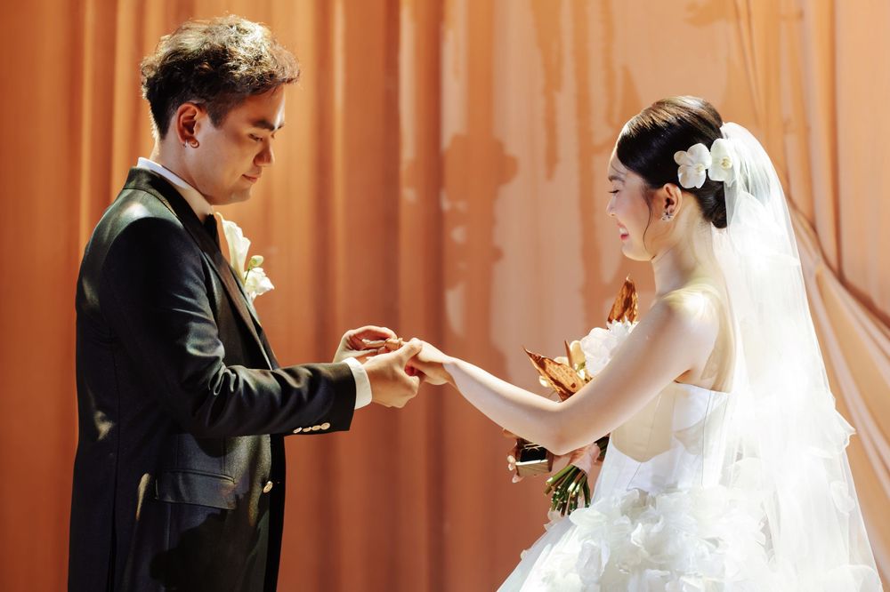 Groom in black tuxedo places wedding ring on brides finger during elegant indoor ceremony.