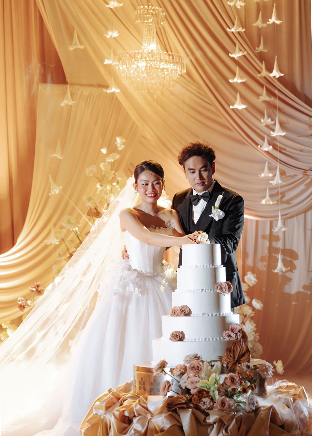 Bride and groom cutting a tall white wedding cake before golden draped backdrop with paper cranes.