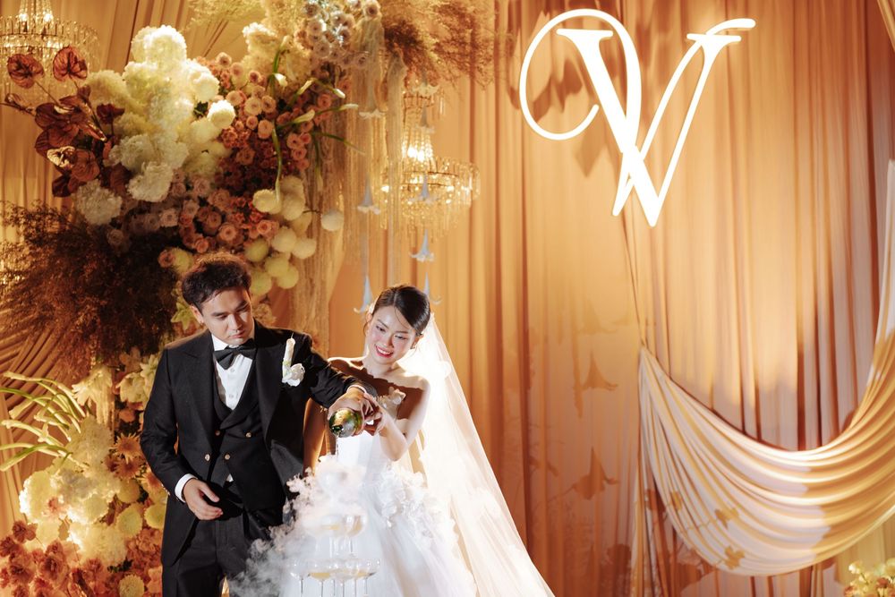 Bride and groom share a tender moment before a golden floral backdrop with monogram V decor.