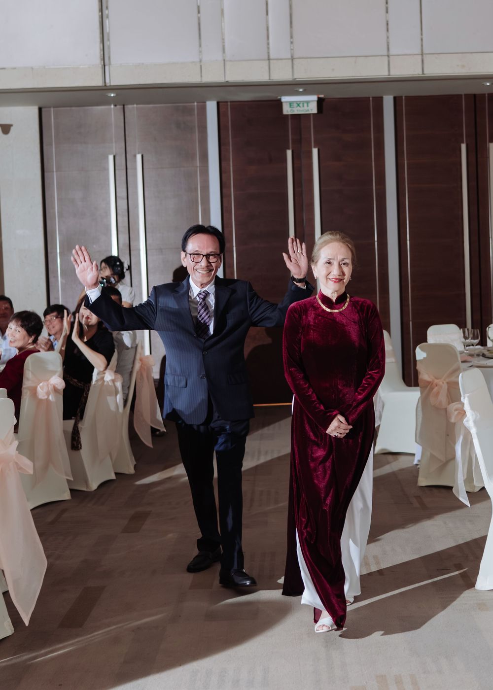 Elegant couple in formal attire waving to guests at a wedding reception banquet hall.