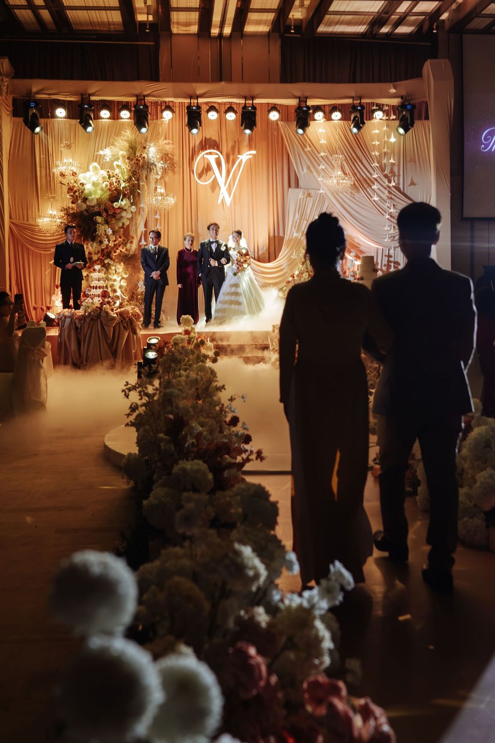 Silhouettes of guests watching bride and groom on a floral-lined illuminated wedding ceremony stage.