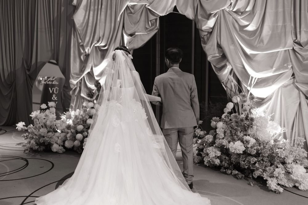 Black and white photo of bride and groom walking toward floral arch at elegant wedding ceremony.