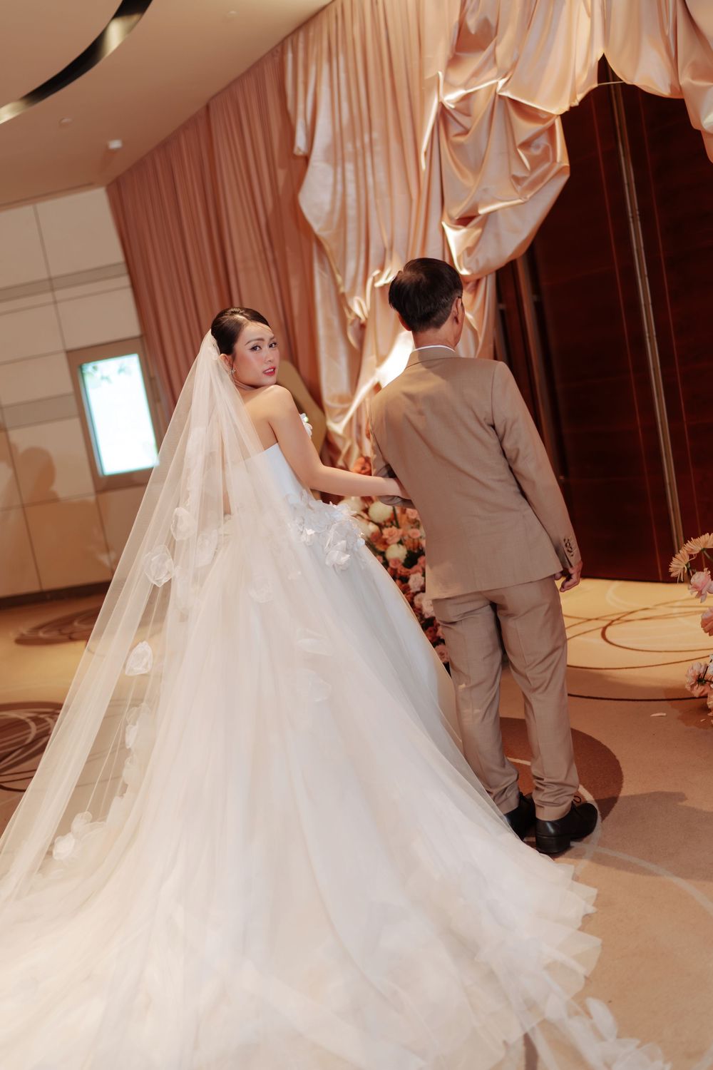 Bride in white ball gown with long veil holds groom's hand walking toward wedding ceremony altar.