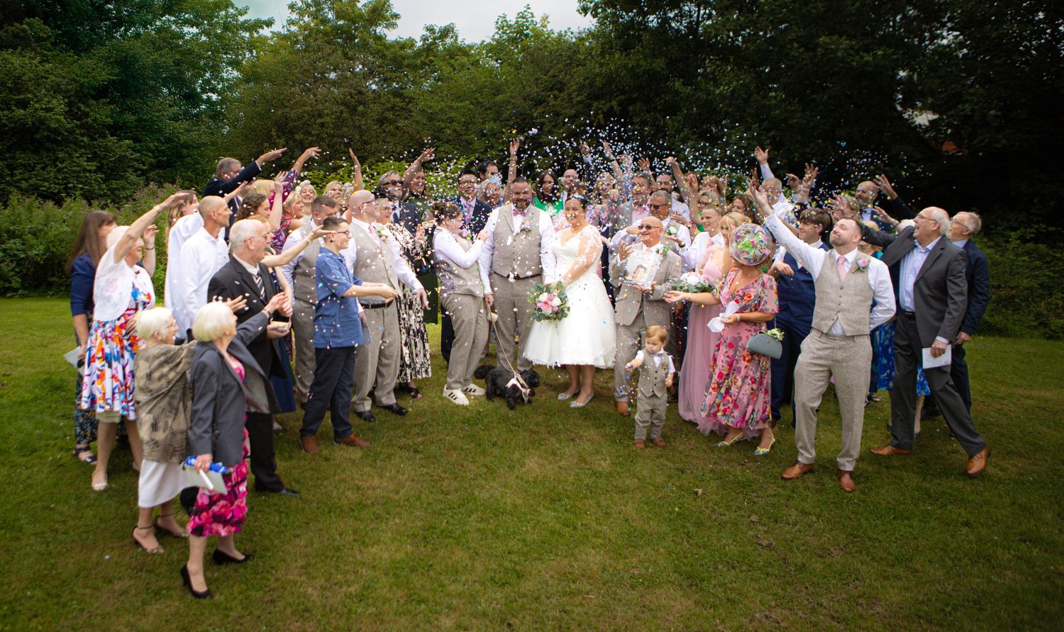A large group of wedding guests gather in a relaxed outdoor garden setting for a celebratory photo.