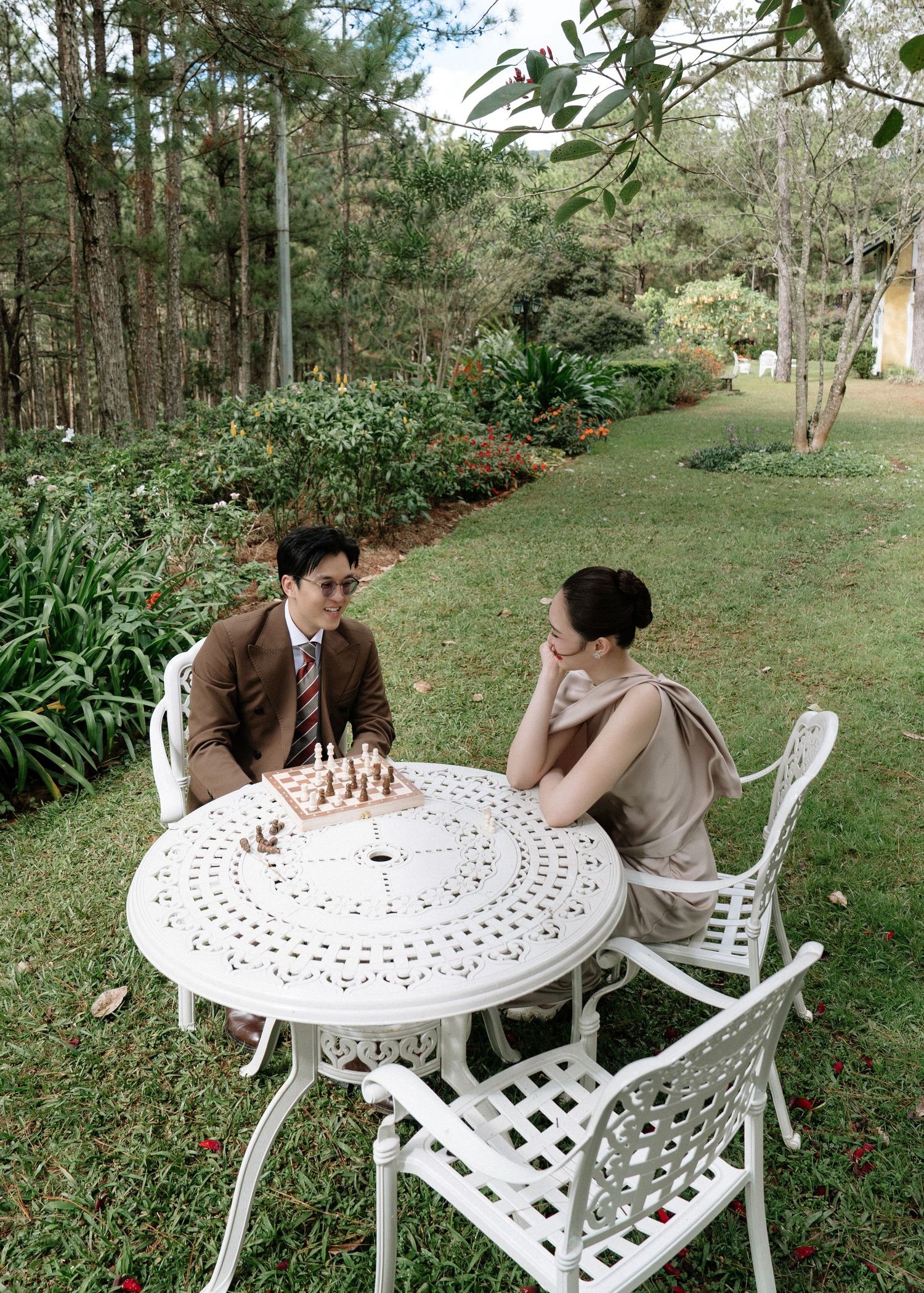Couple seated at a white ornate garden table playing chess surrounded by lush greenery.