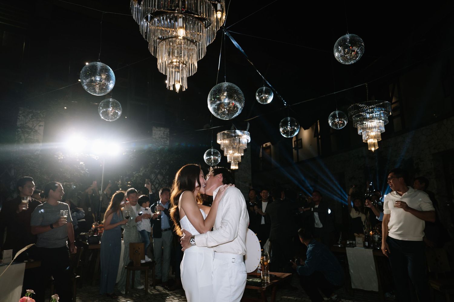 Bride and groom sharing a romantic kiss on the dance floor under sparkling chandeliers and disco balls at their wedding reception.