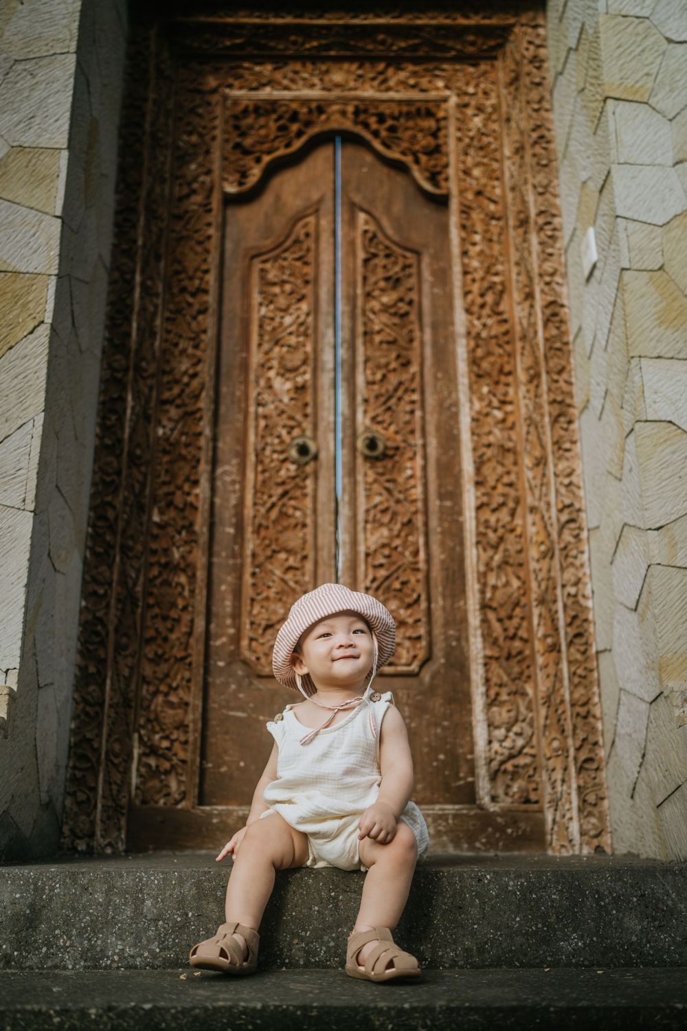 A small child sitting by an ornately carved stone wall with traditional Balinese designs.