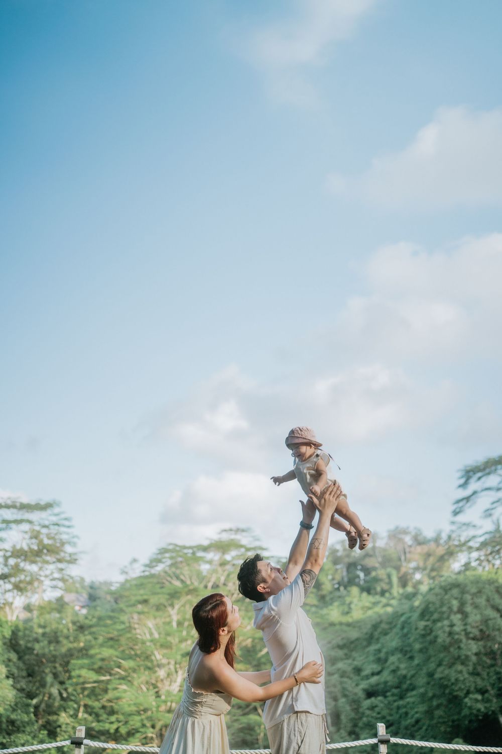 Parents playing with their child against a backdrop of tropical trees and blue sky.