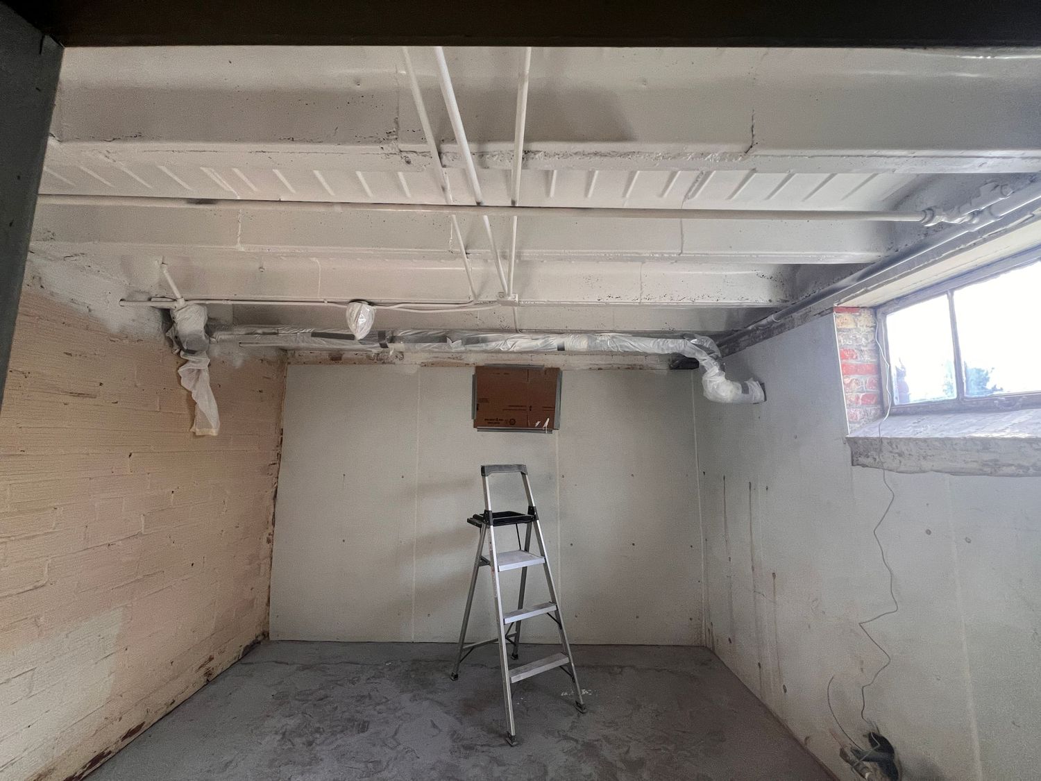 Empty basement space with exposed pipes and ductwork on white ceiling.