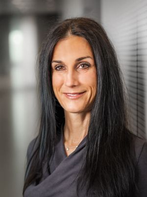 Professional headshot of a person with long dark hair wearing a black top against a blurred office background.