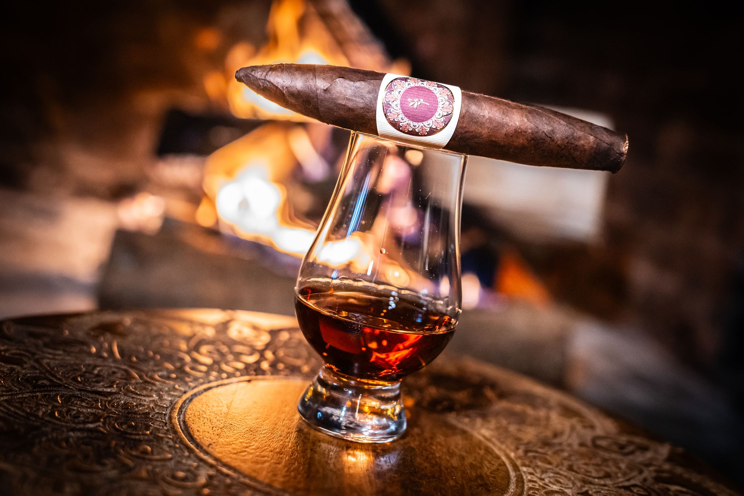 A burning cigar rests on an ornate ashtray next to a glass of whiskey with soft ambient lighting.