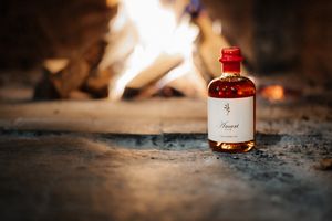 A bottle of amber spirits sits on stone before a glowing fireplace, warm flames illuminating the elegant label.