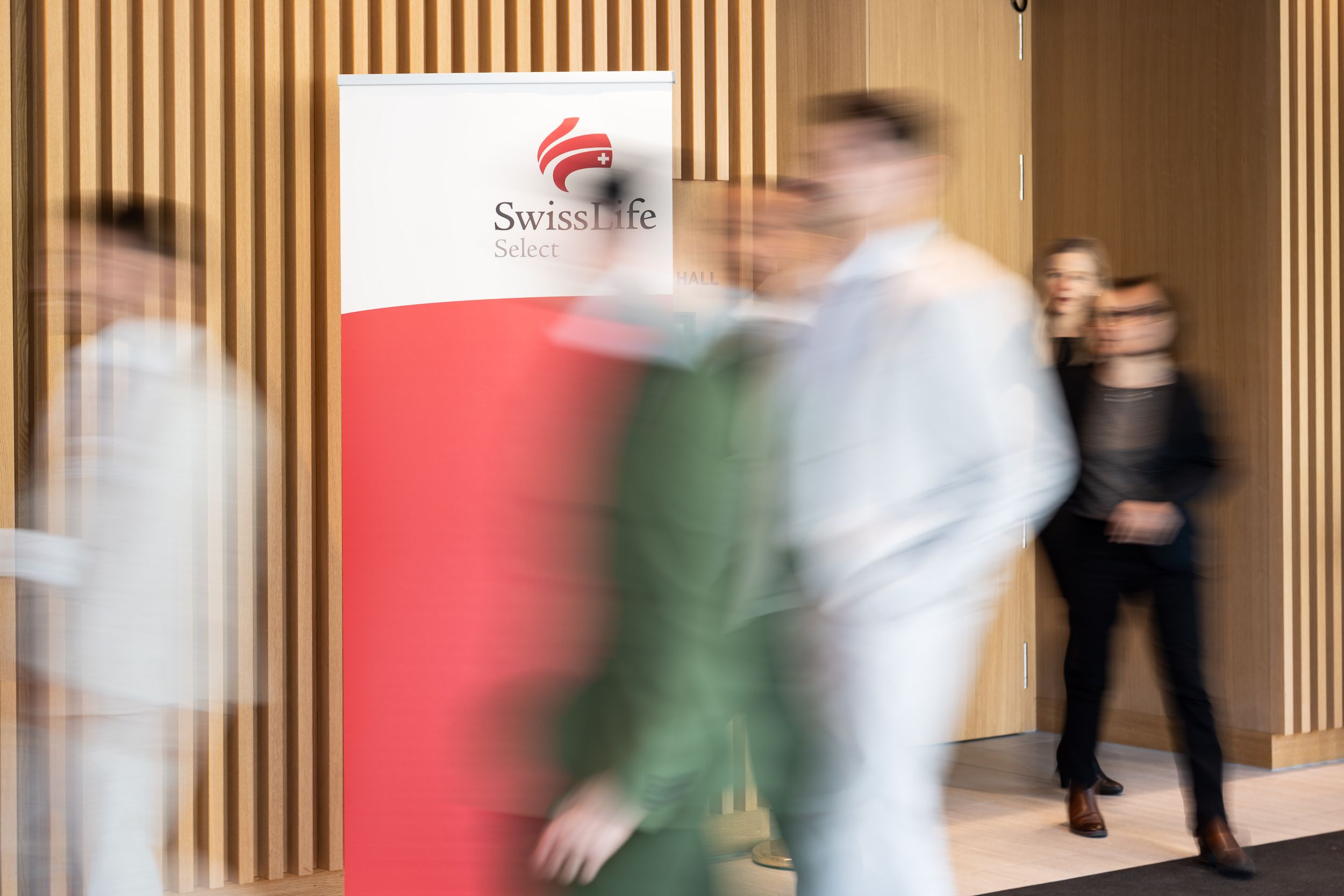 Motion blur of people walking in a modern office hallway with wooden slat walls.