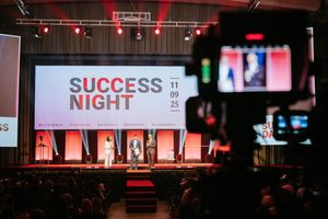 Conference stage with red lit backdrop and white SUCCESS text displayed above dark seated audience.
