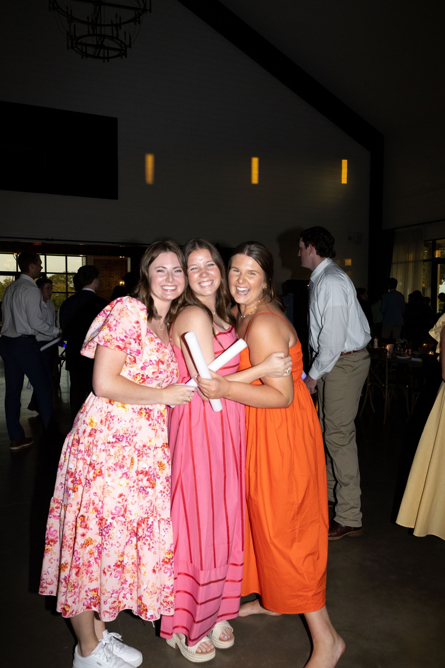 Three friends pose together at an event wearing pink, coral and orange dresses in a dimly lit reception hall.
