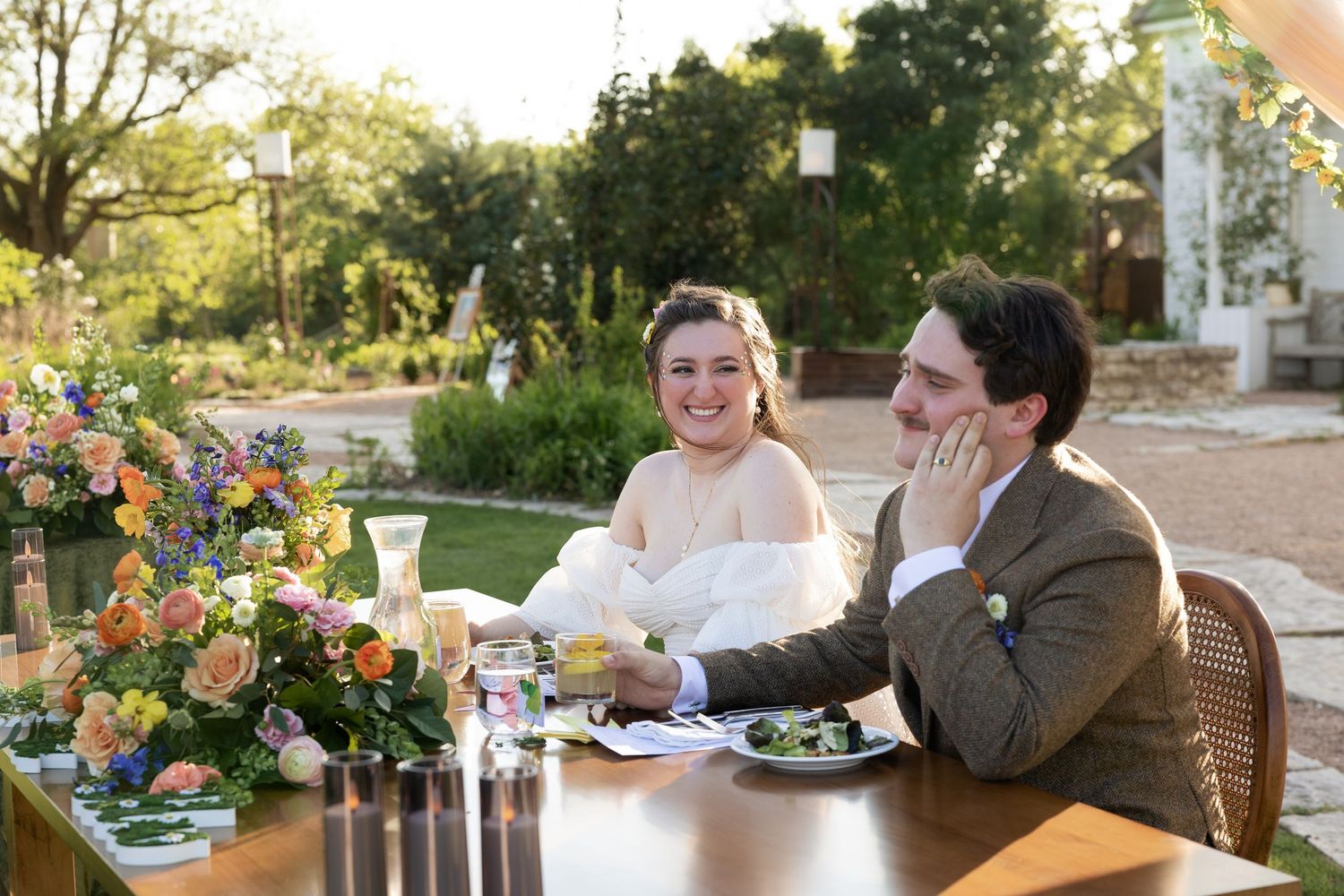 People gather to eat at an outdoor garden party with colorful floral arrangements and rustic table settings.