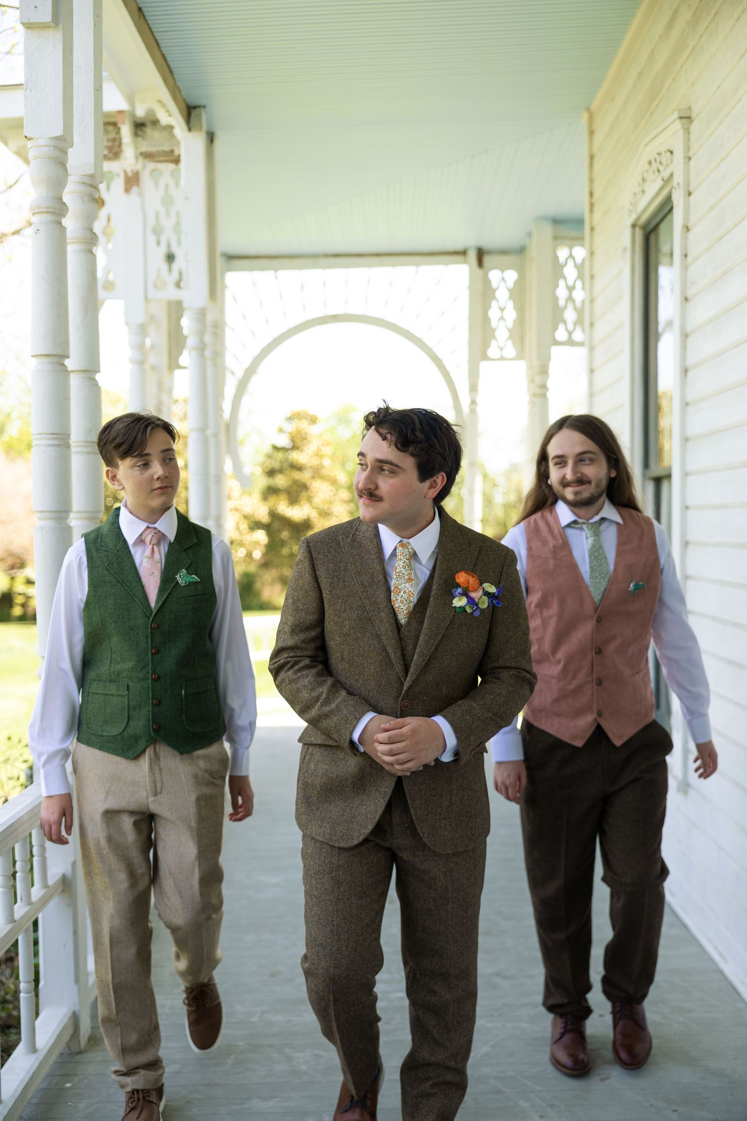 A row of groomsmen in earth-toned suits walk together along a covered porch of a white farmhouse.