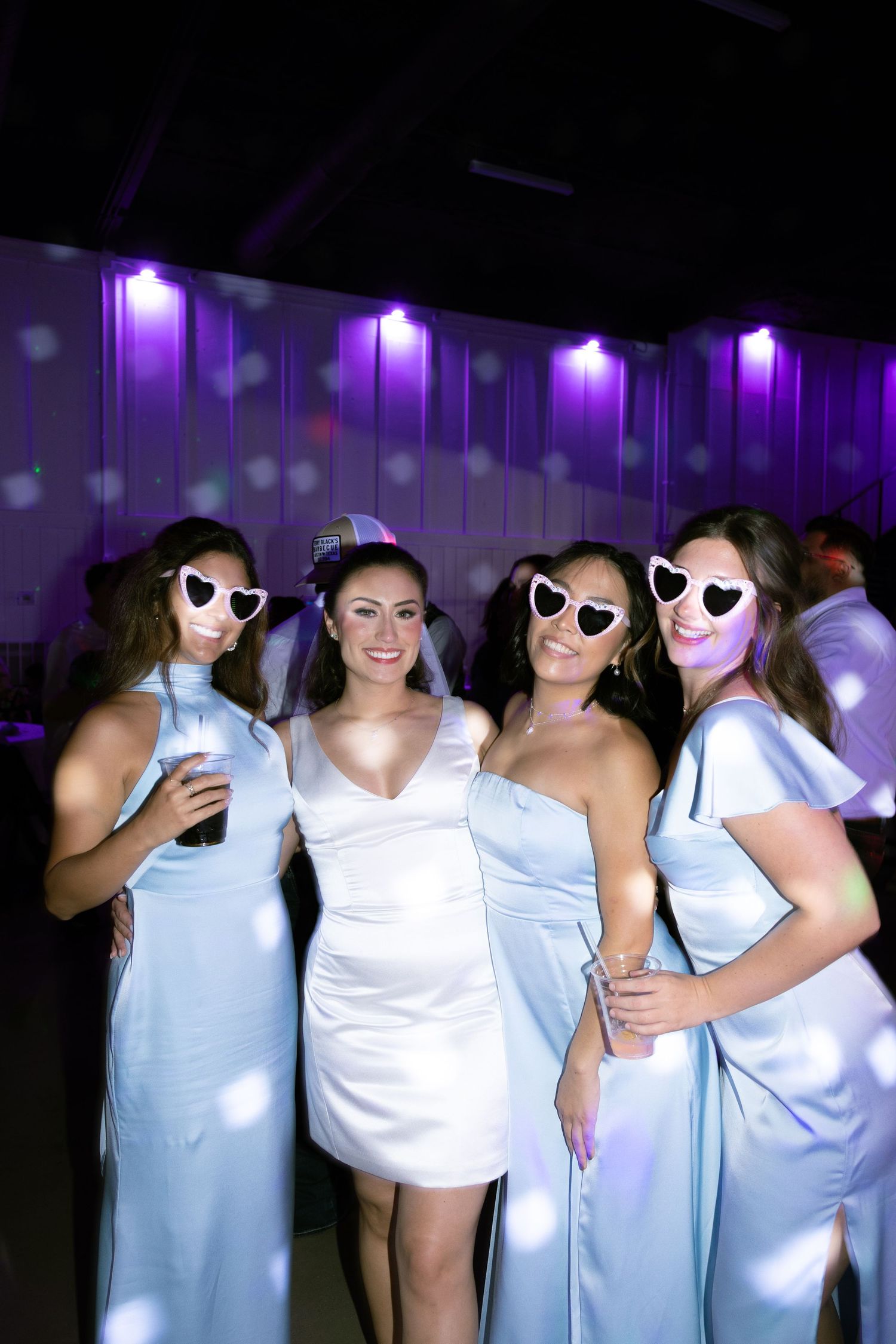 Party guests wearing white outfits and heart-shaped sunglasses pose together under purple lighting at a nighttime event.