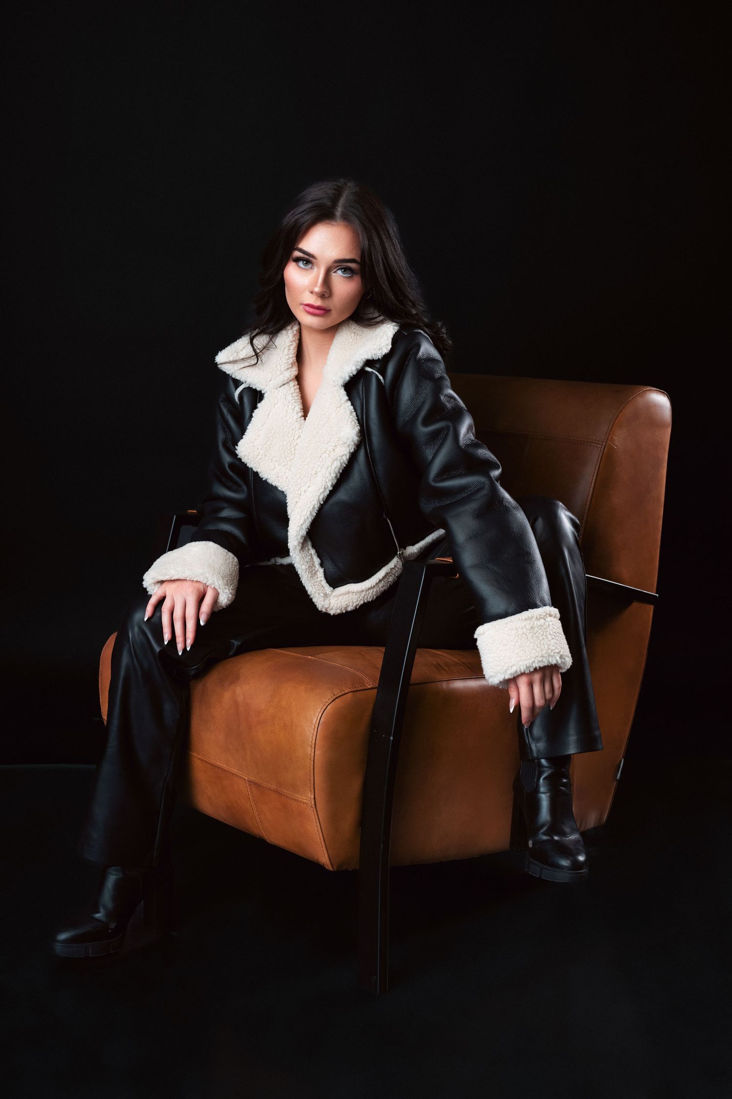 Fashionable winter outfit with black leather shearling coat and boots styled on brown leather armchair against dark backdrop.