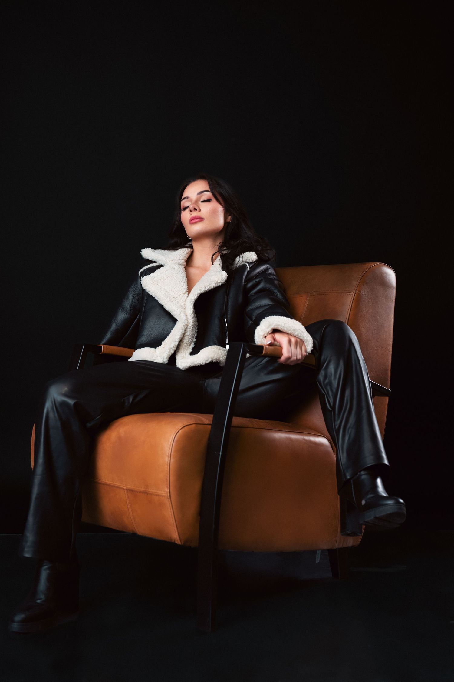 A figure in a black leather jacket and white collar reclines dramatically on a brown leather couch against a dark background.