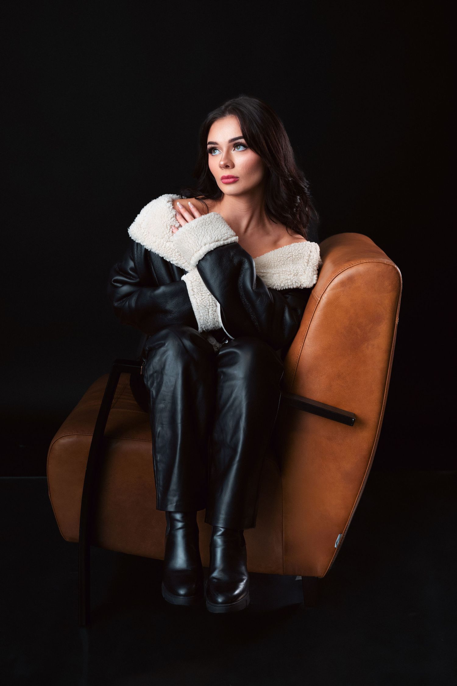 Fashionable model in leather pants and white sweater poses on brown leather armchair against dark background.