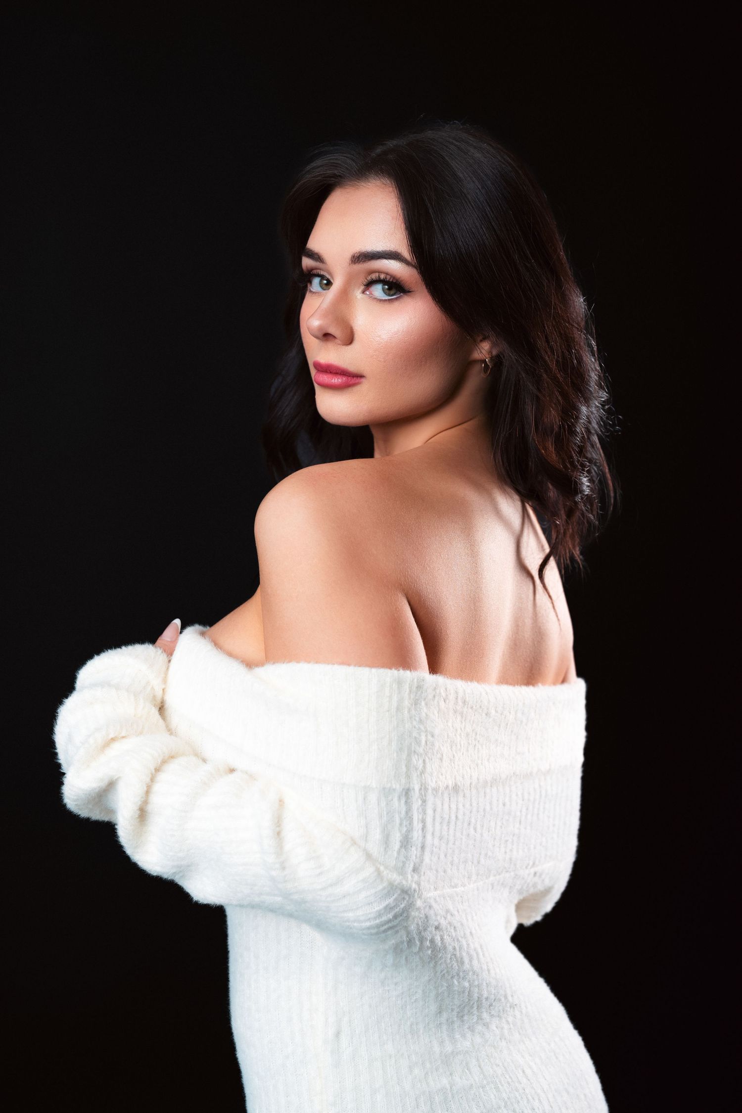 Model in white off-shoulder sweater poses gracefully against dark background showing elegant glamour style.