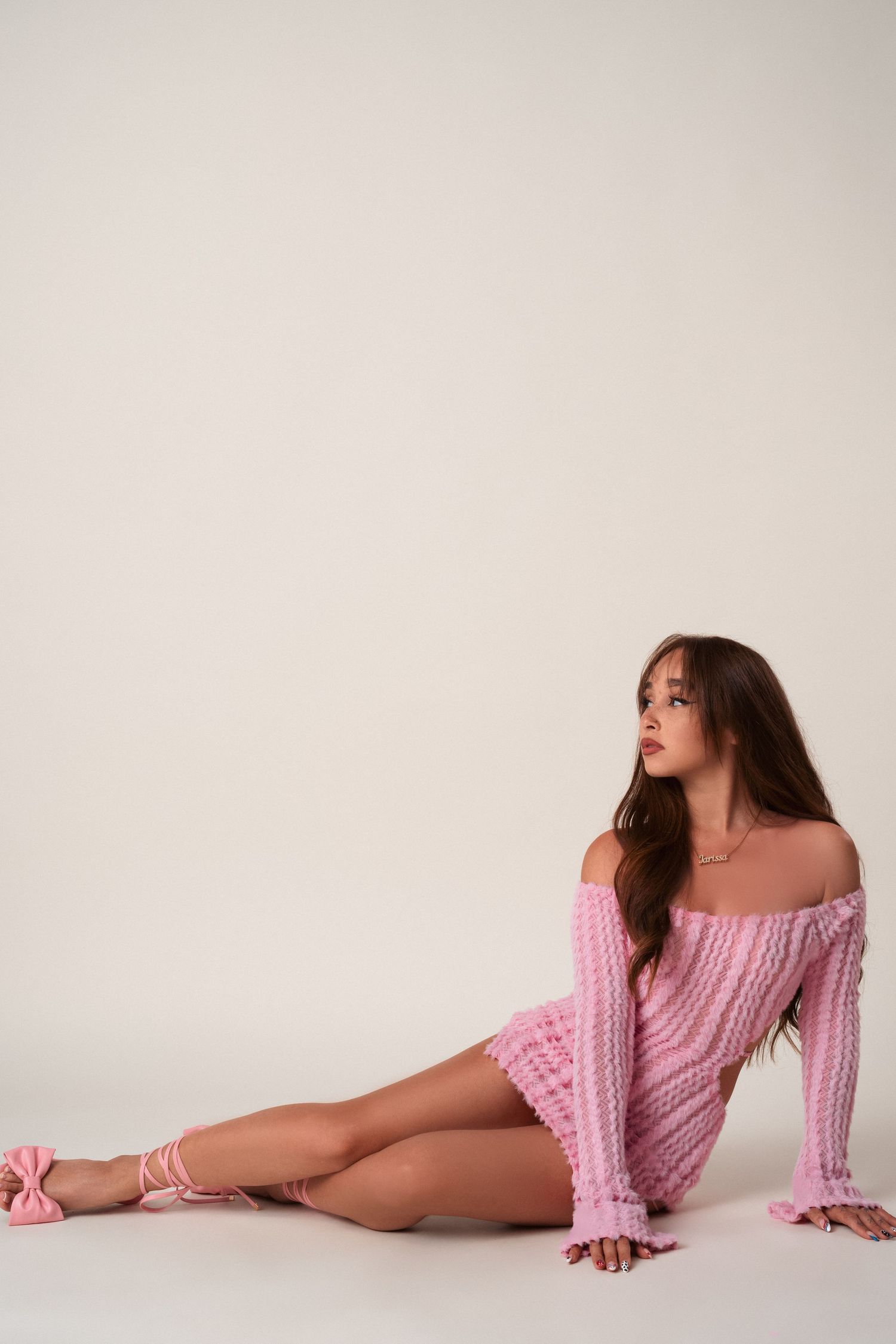 A person in a cozy pink off-shoulder sweater sits on a white floor in an artistic pose.