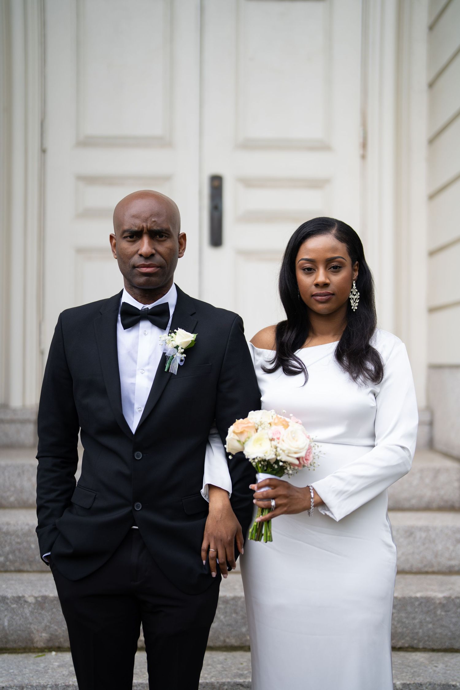 A wedding couple in elegant attire poses for a series of formal portraits against a white exterior wall.