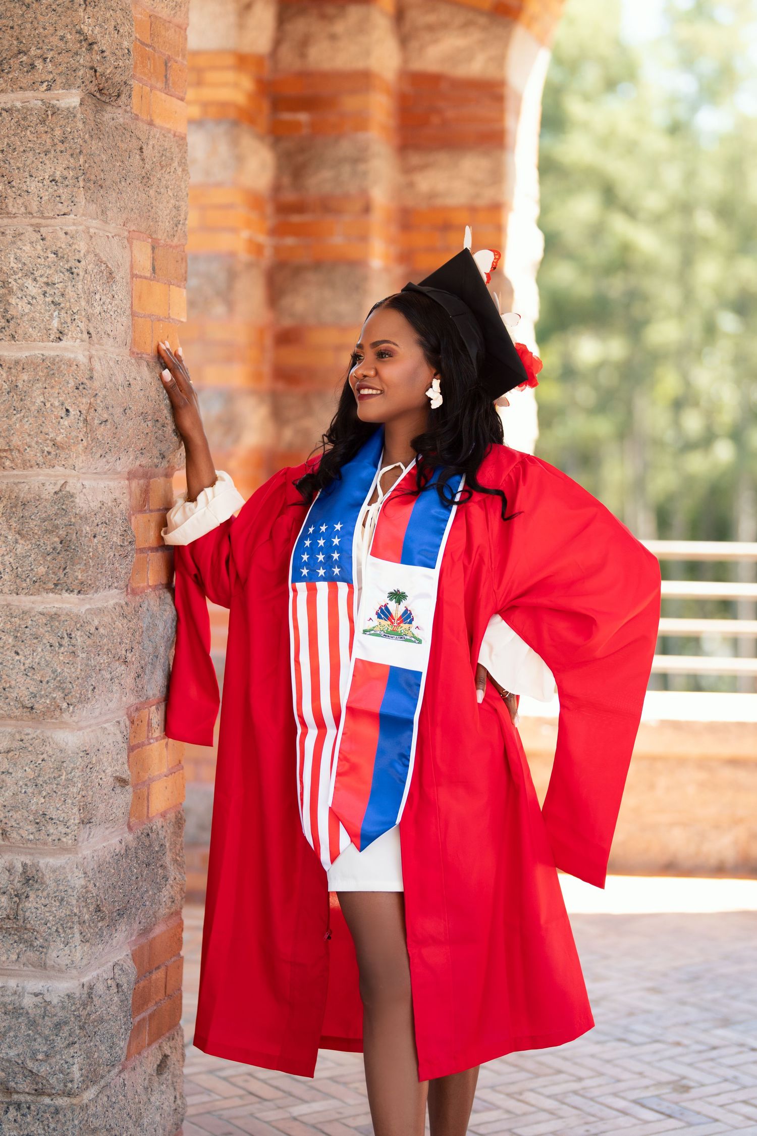 Student in red graduation gown and Haitian-American flag shirt smiles and poses during commencement celebration.