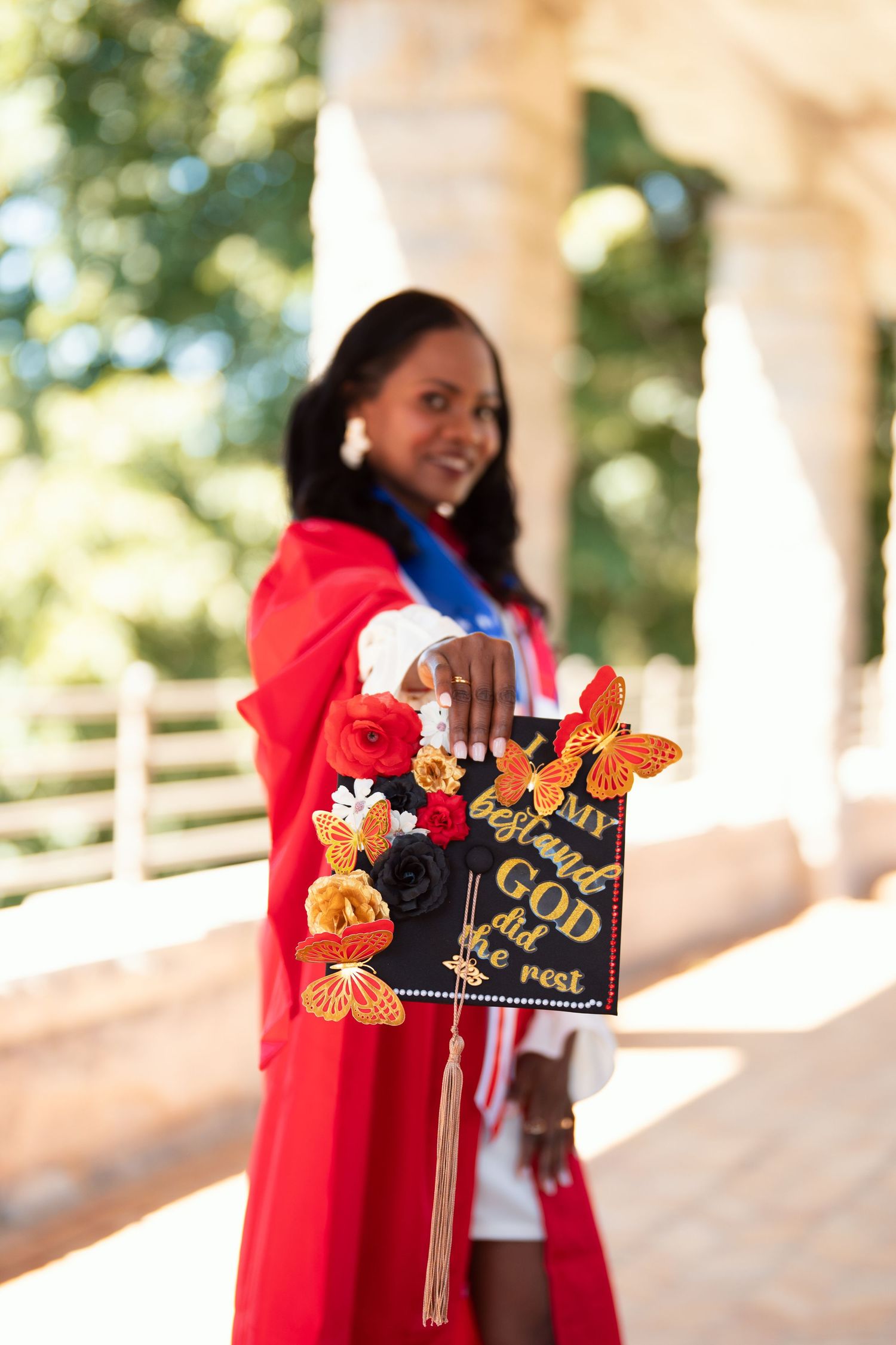 Graduate in a red academic gown holding up decorated graduation cap with butterfly designs in outdoor campus setting.
