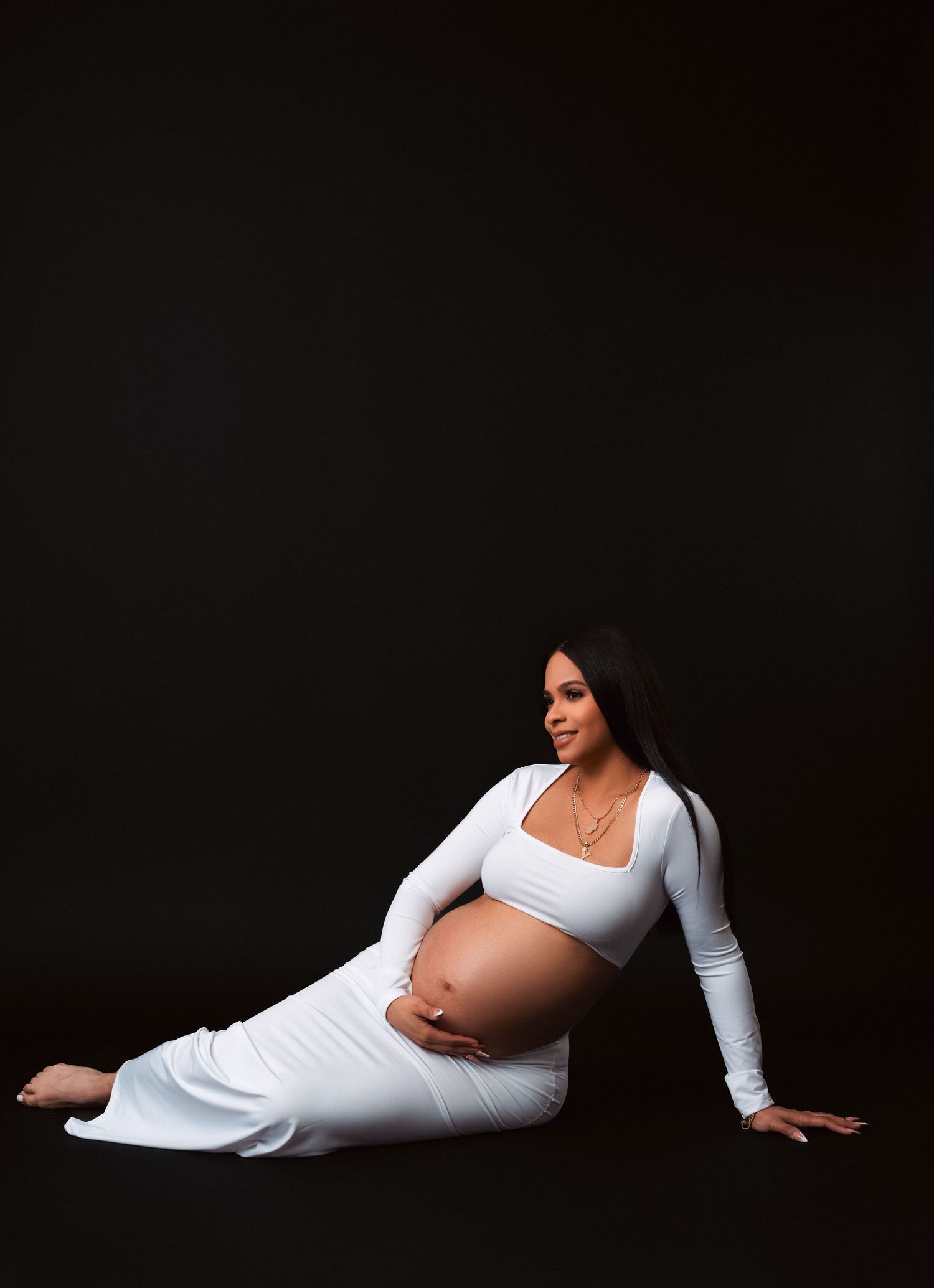 A maternity photo against dark background shows a pregnant woman in white attire posing elegantly on the floor.