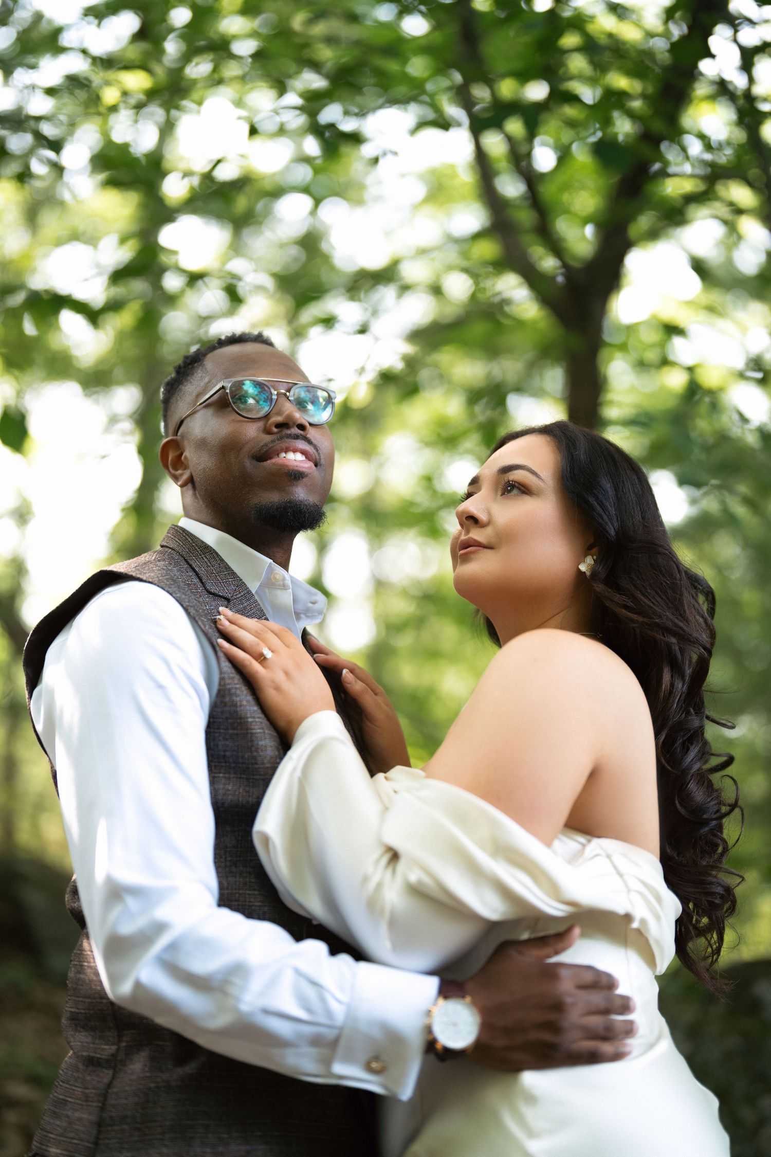 A romantic couple embraces and dances outdoors in a natural garden setting with sunlit trees in the background.