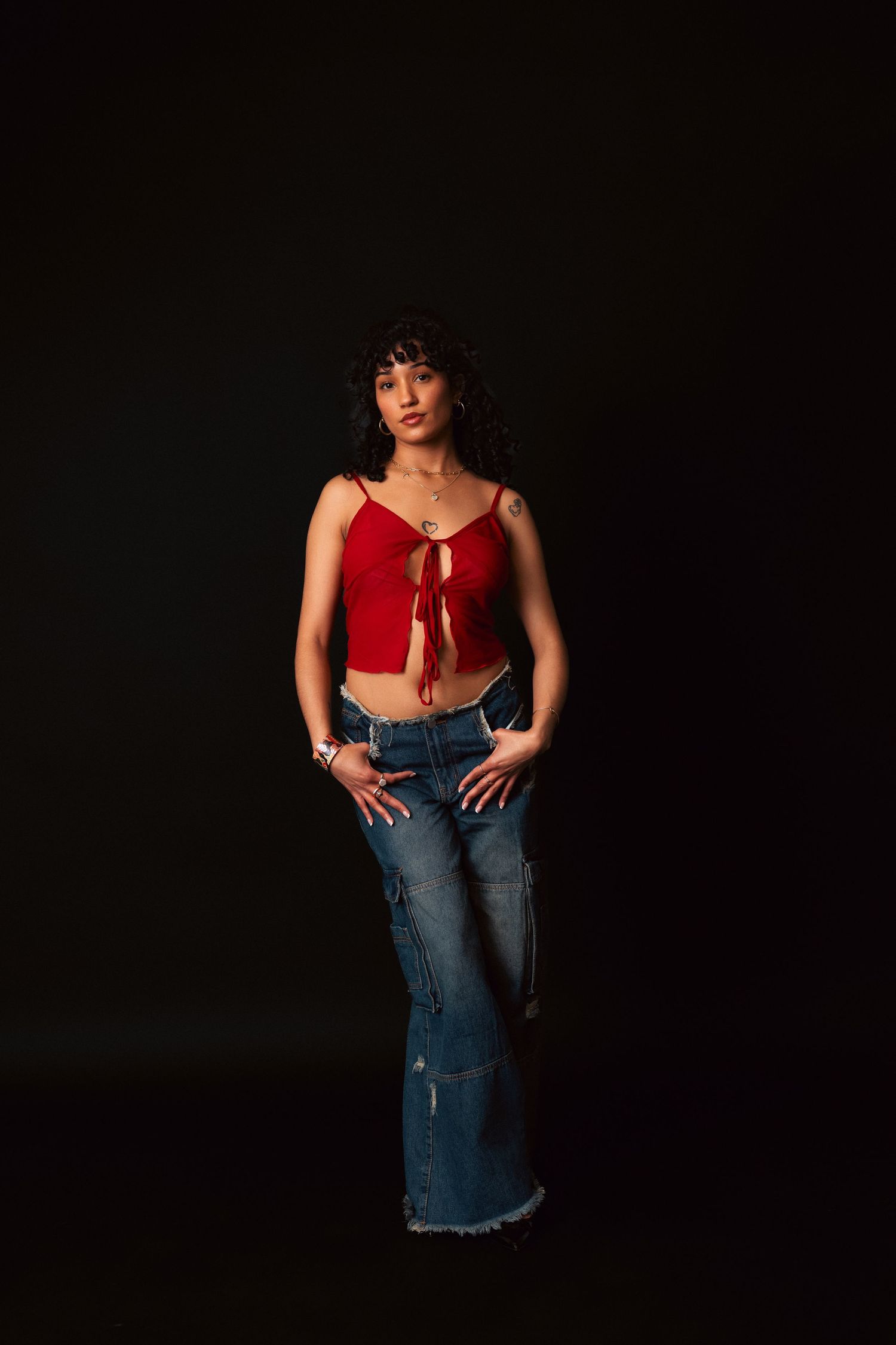 Woman poses against dark background wearing red crop top and distressed flared jeans in casual fashion portrait.