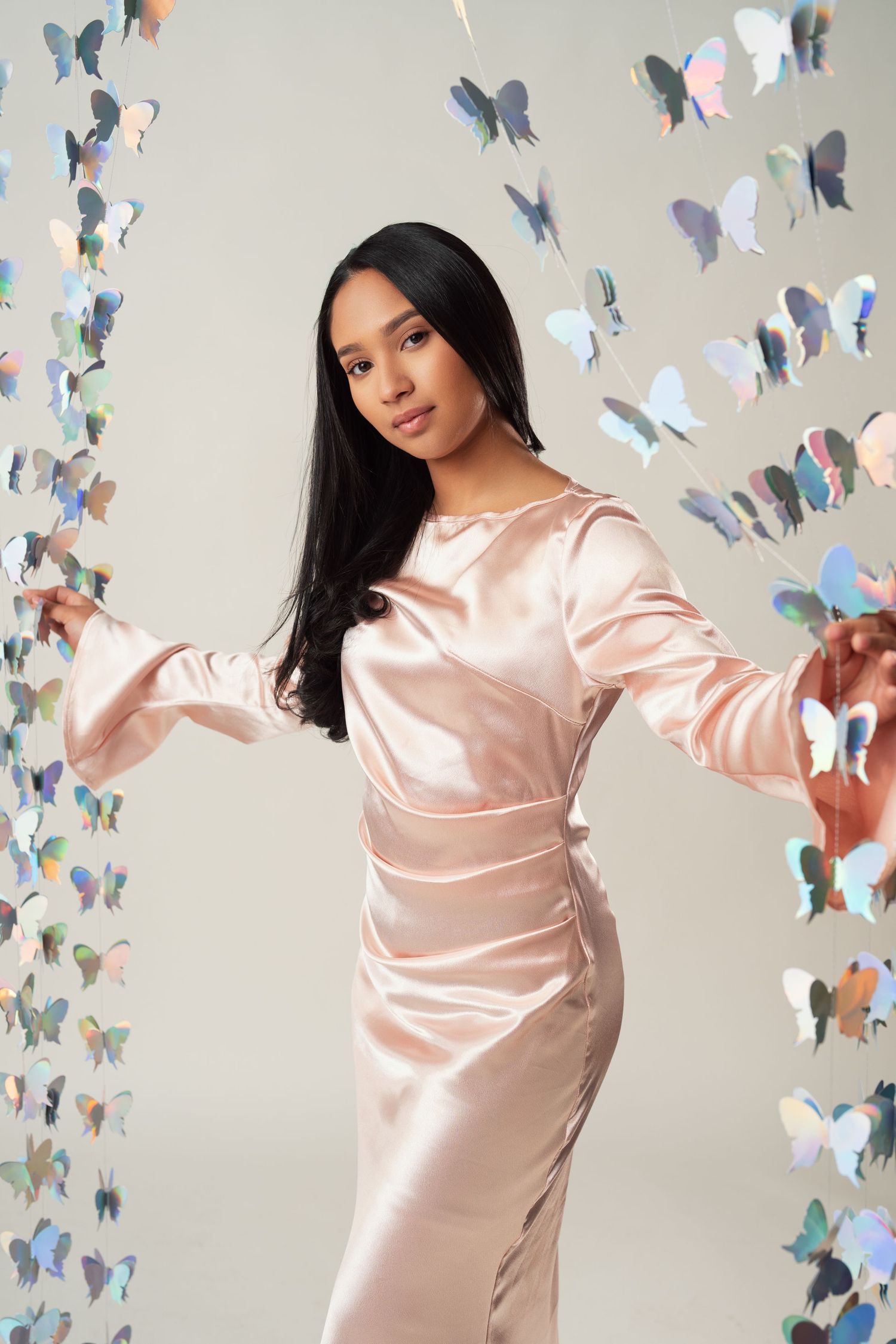 Model in pink silk dress poses elegantly against wall decorated with iridescent butterfly decorations.
