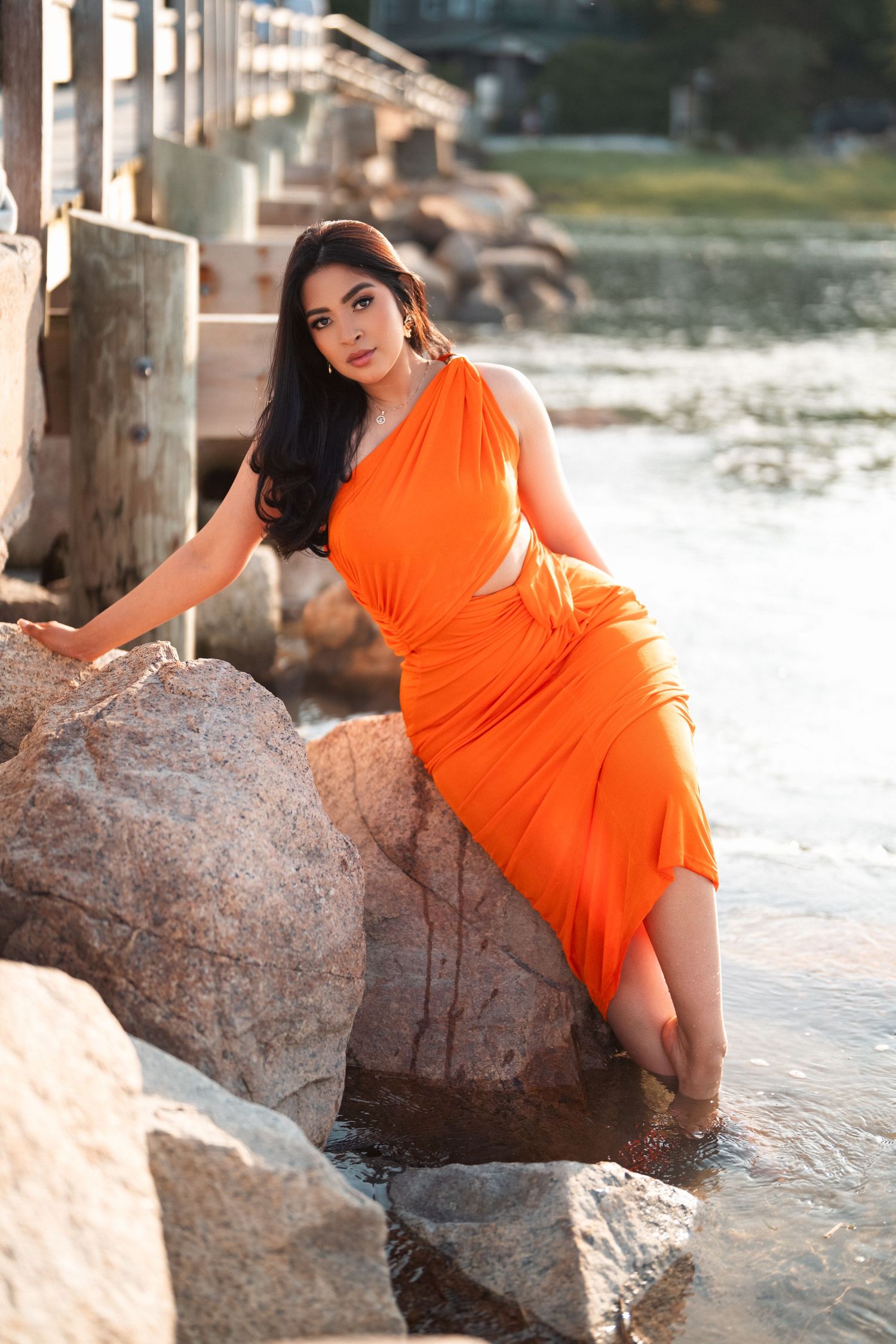 Elegant orange one-shoulder dress flows gracefully by the rocky shoreline at sunset with water and pier in background.