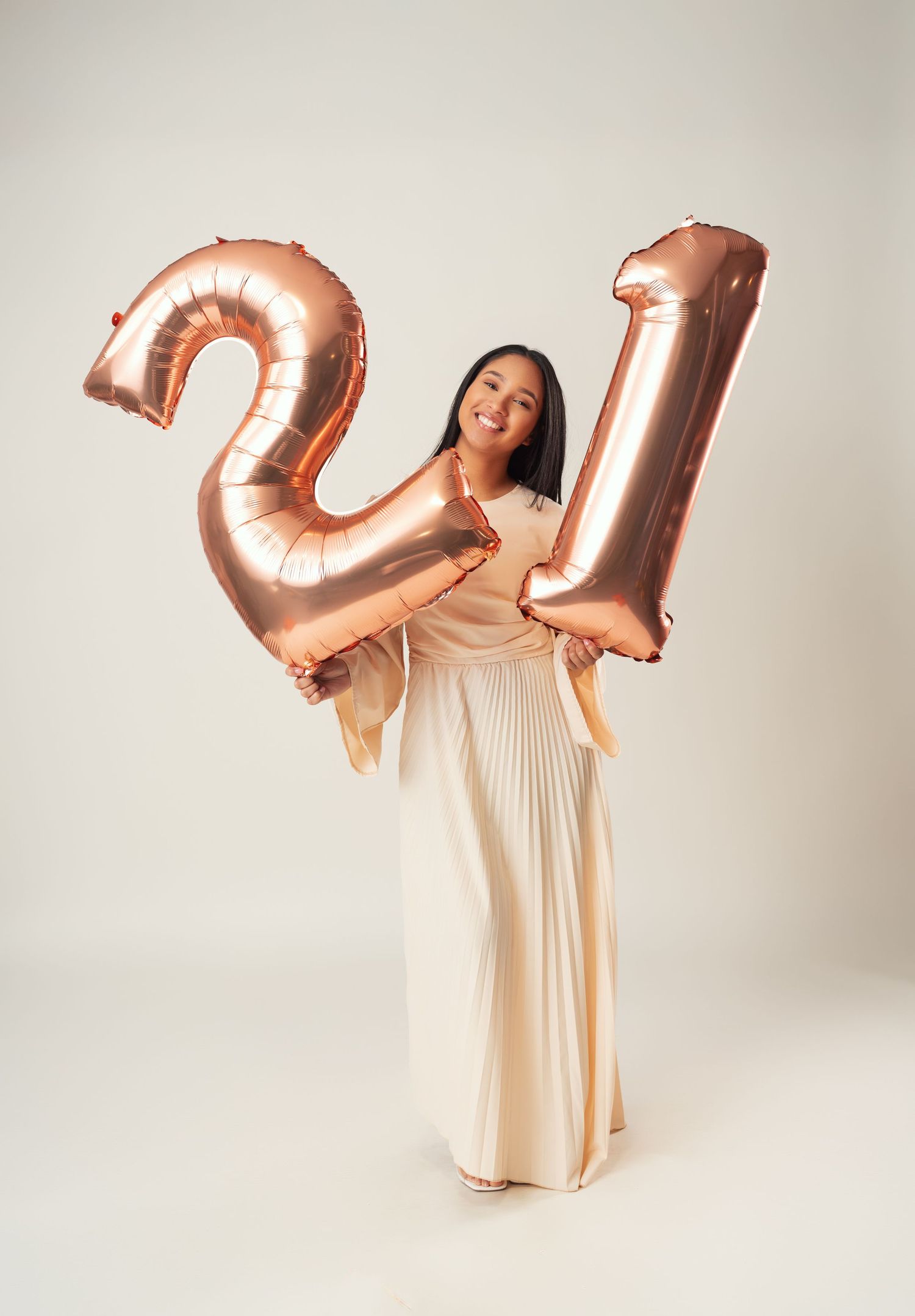 Person in flowing cream dress poses with rose gold number balloons in minimalist studio setting.
