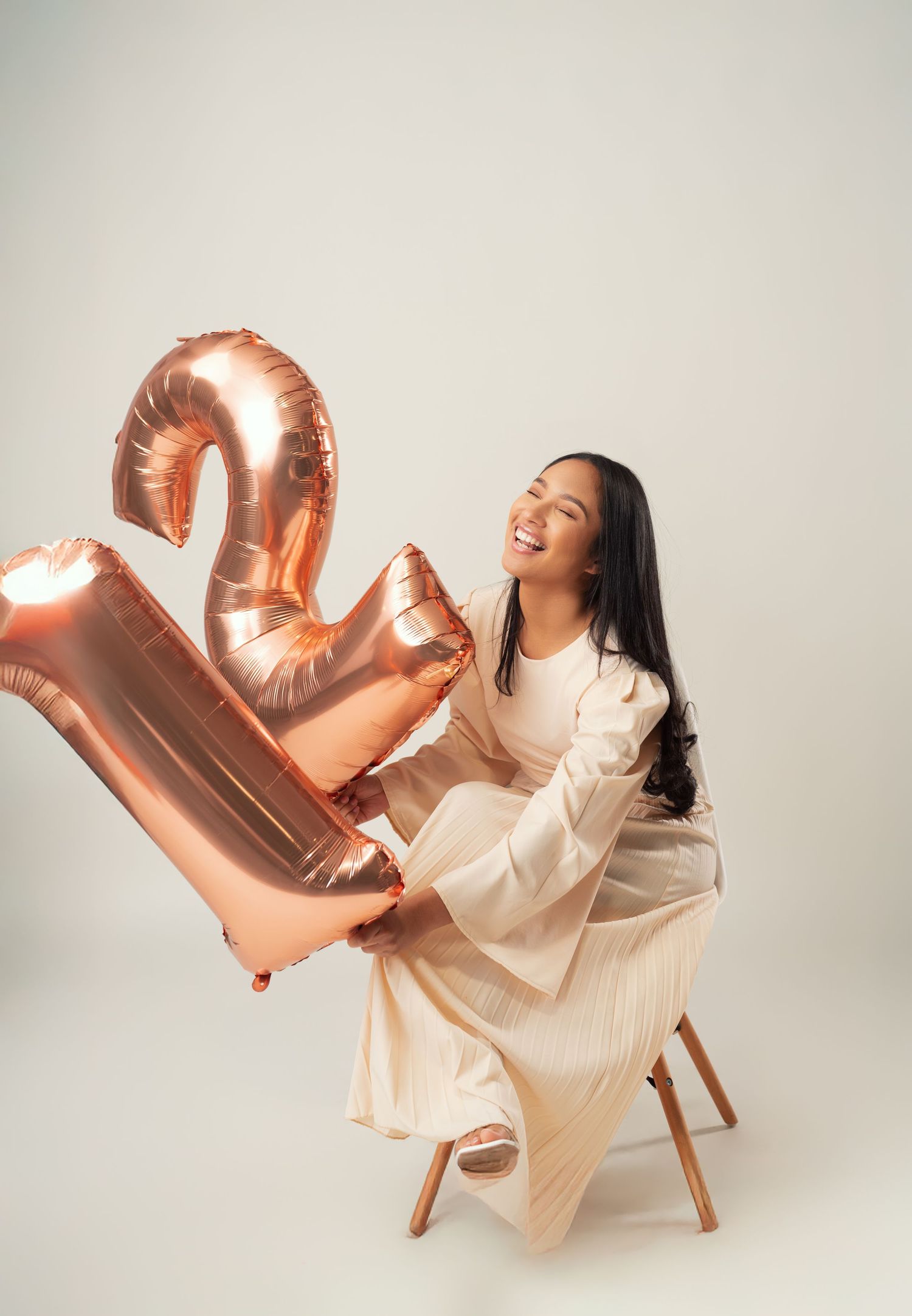 Person in white lounges playfully on chair with rose gold balloon numbers in minimalist studio setting.