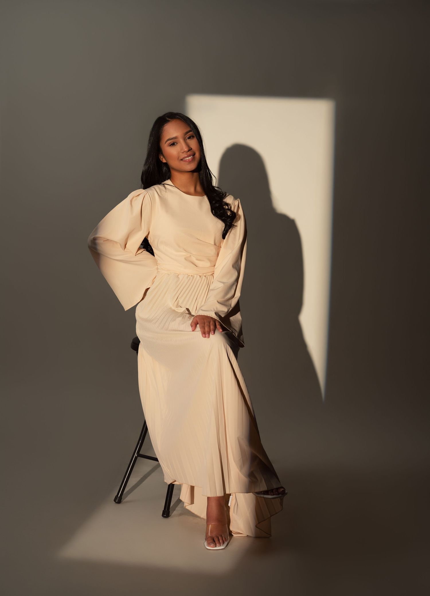 Model in cream-colored wrap dress poses against dark backdrop with dramatic shadows for fashion photoshoot.