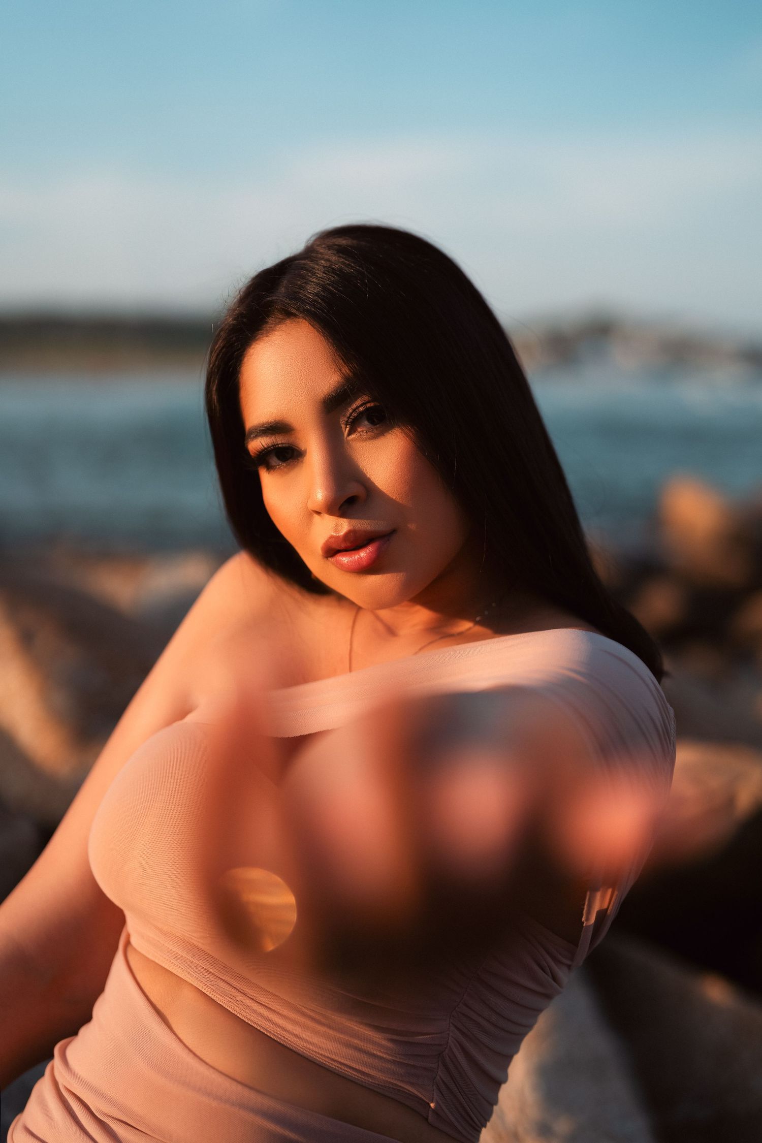 Portrait of a woman in a beige top posing outdoors at sunset near the ocean with warm lighting and blurred background.