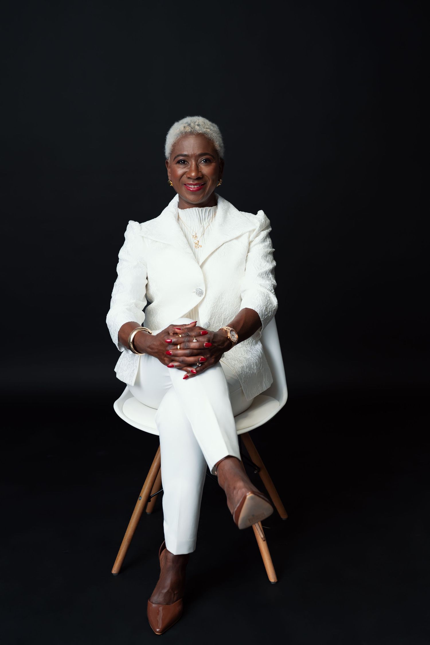 A figure in an all-white suit sits posed in a wooden chair against a dark background in varied stances.