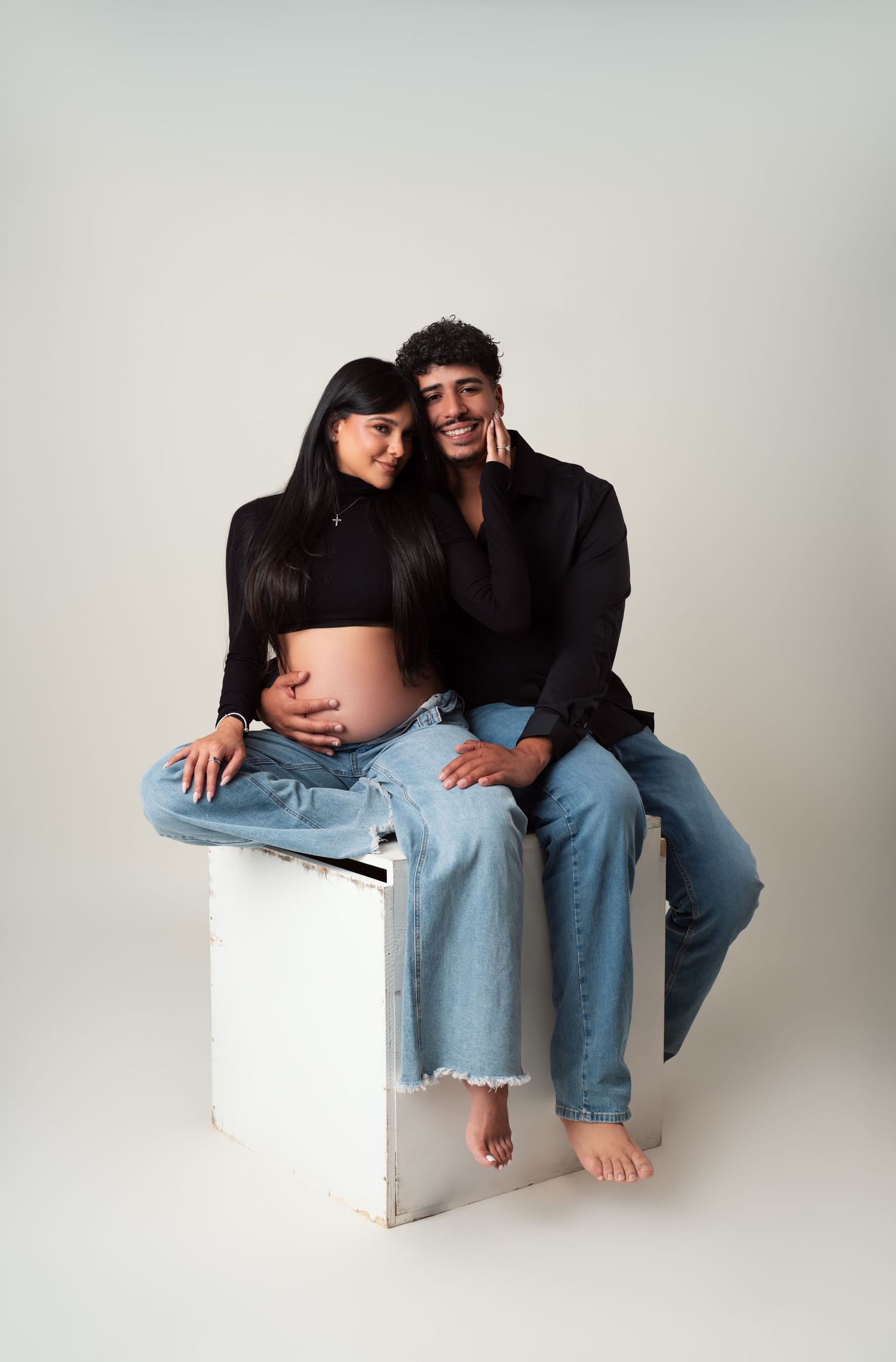 Two people in casual attire sit intimately together on a white platform in a minimalist studio setting.